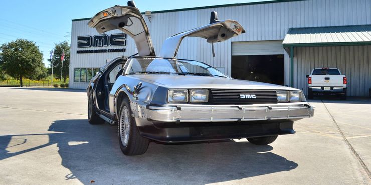 Stainless steel DeLorean DMC-12 car with gullwing doors (doors open straight up). The car is sitting in front of a DMC shop.
